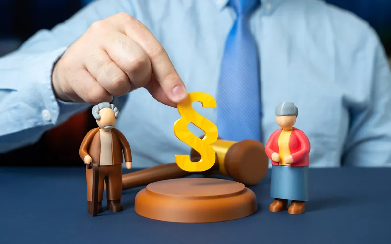 Toy figures of a man and woman beside a gavel and legal symbol, held by a man in a blue shirt and tie, on a blue surface.